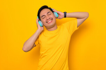 Fototapeta na wymiar Relaxed young Asian man in casual t-shirt listening music in headphones isolated on yellow background. People emotions lifestyle concept