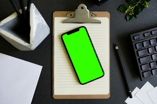 Top view Smartphone with greean screen is on top of office desk table with keyboard,pen stand,paper etc