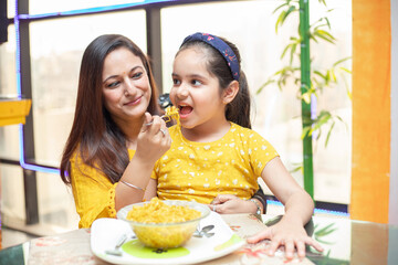 Young Indian mother feed her little daughter noodles breakfast at table, Little girl child having...