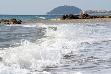 Beach on the Italian Riviera with sun, sea and stones in the sea water. Rough Sea and Foamy Waves on Shore at Mediterranean Coast in Sunrise