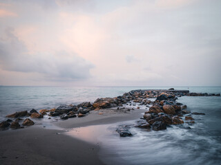 Obraz premium Beach on the Italian Riviera with sun, sea and stones in the sea water. Rough Sea and Foamy Waves on Shore at Mediterranean Coast in Sunrise