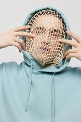 a close vertical, conceptual studio photograph of a strange man in a light blue hoodie covering his face with his hands, standing on a light background with a hood on his head