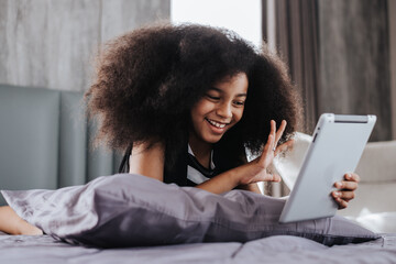 African girl playing digital tablet on bed in bedroom. African girl using tablet to video call talking with friends.