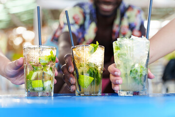 Best friends toasting cocktails. Multiethnic young people drinking long drinks at beach bar. Shallow depth of field with focus on hands holding iced mojitos. Close up shot, focus on the center glass.