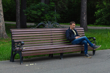Lonely adult man sits alone on bench in park, holds smartphone and uses it, selective focus Outdoors