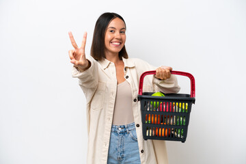 Obraz na płótnie Canvas Young woman holding a shopping basket full of food isolated on white background smiling and showing victory sign