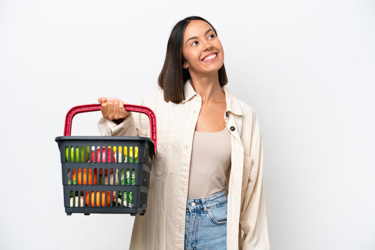 Young woman holding a shopping basket full of food isolated on white background thinking an idea while looking up
