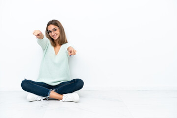 Young caucasian woman sitting on the floor isolated on white background points finger at you while smiling
