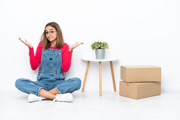 Young caucasian woman sitting on the floor among boxes having doubts while raising hands