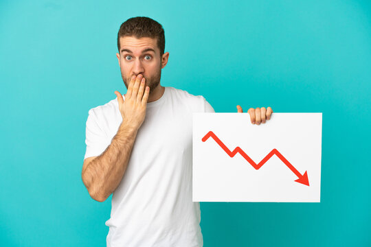 Handsome blonde man over isolated blue background holding a sign with a decreasing statistics arrow symbol with surprised expression