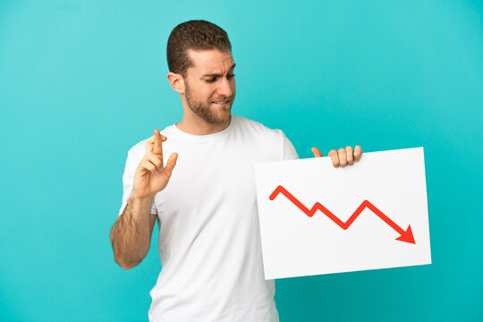 Handsome blonde man over isolated blue background holding a sign with a decreasing statistics arrow symbol with fingers crossing