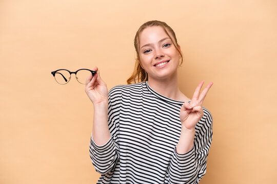 Young caucasian girl with glasses isolated on beige background smiling and showing victory sign