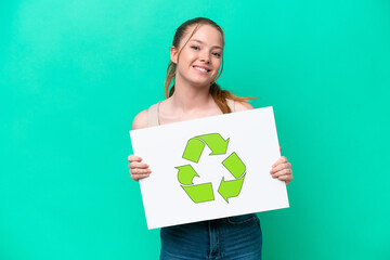 Young caucasian woman isolated on green background holding a placard with recycle icon with happy expression