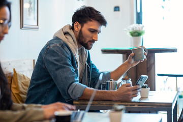 Handsome young man using his smartphone while drinking a cup o coffee in a eco bar.