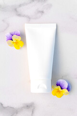 Mockup of white squeeze bottle cosmetic cream tube for branding of medicine or cosmetics - shampoo, gel, skincare. Moisturizer blank bottle and pansy flowers on marble background. Top view.