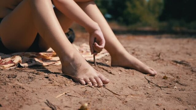 Bored Naked Child Sits on a Coverlet, Draws with a Twig in Sand on Beach. Close-up of bare legs, arms, a lonely, tired boy drawing on the ground of boredom. Heat, outdoor recreation, summer. Nature.