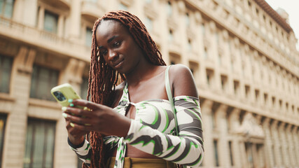 Smiling gorgeous woman with African braids wearing top stands on the sidewalk next to the road and uses mobile phone. Stylish girl writes message on smartphone