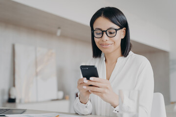 Fototapeta premium Businesswoman in glasses using phone mobile apps in office, reads good news message, chatting online