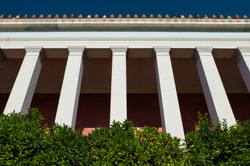 Landscape picture of a symmetrical row of columns of an old greek building in Athens, Greece. Bushes in the foreground and decoration elements on the roof corner.