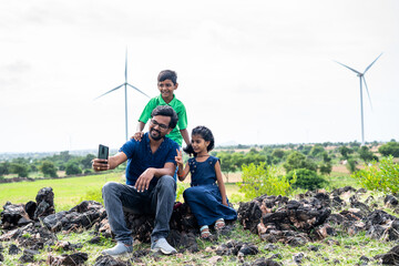 Happy father and kids taking selfie on mobile phone in front of wind turbines or power plant at...