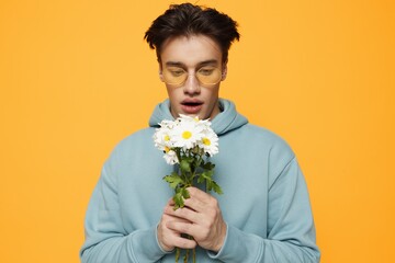 a close portrait of a funny man standing on an orange background in a light blue hoodie, wearing yellow glasses, holding a bouquet of white daisies looking at him