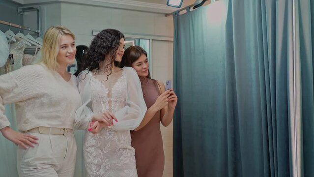 Attractive bride with curly hair in lace wedding dress taking photo with friends in bridal salon. Slow motion.