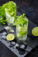 Mojito cocktail, lime slices, mint and ice on a dark background. Cocktail mojito, ice tongs and cocktail spoon on slate board. Top view