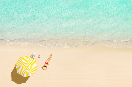 Top view of woman in bikini and straw hat lying and sunbathes near sunbed, lounge, flip flops, yellow umbrella on tropical Seychelles sea sand beach. Aerial, drone view