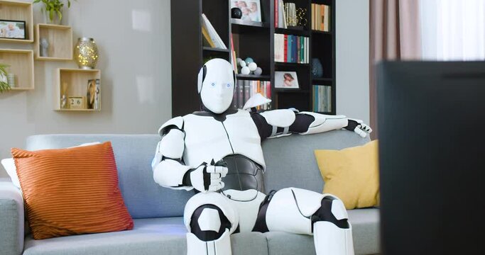 Humanoid droid sitting on couch and using remote control for switching channels while watching TV. Concept of future technology and domestic leisure time.