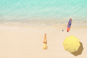 Top view of woman in bikini and hat lying near umbrella and sunbathes on tropical Seychelles sea sand beach with SUP board for surfing. Aerial, drone view