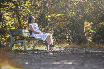 Beautiful woman sitting in a city park on a wooden bench. The woman is wearing long white hair and a straw hat laid side by side. It is a beautiful sunny day and there are trees and nature around.