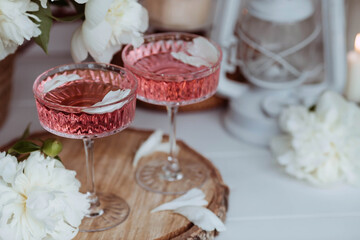 A bouquet of white peonies, flower petals, candles and a fashionable glass for dessert and champagne with a pink drink inside. The concept of a party and a holiday.