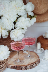 A bouquet of white peonies, flower petals, candles and a fashionable glass for dessert and champagne with a pink drink inside. The concept of a party and a holiday.