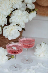 A bouquet of white peonies, flower petals, candles and a fashionable glass for dessert and champagne with a pink drink inside. The concept of a party and a holiday.