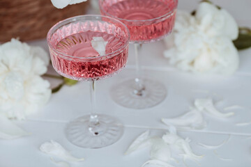 A bouquet of white peonies, flower petals, candles and a fashionable glass for dessert and champagne with a pink drink inside. The concept of a party and a holiday.