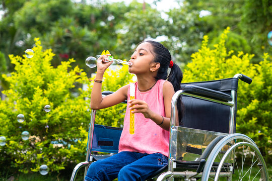cheerful girl kid with disability playing by blowing water bubbles while using wheelchair at park - concept of playful activities, happiness and relaxation.