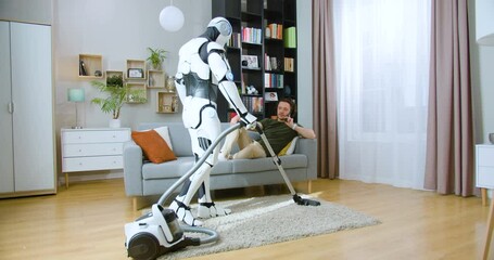 Humanoid robot vacuuming carpet while man in casual wear lying on grey sofa at home. Young guy using modern smartphone to talk while android cleaning apartment.