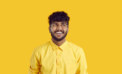 Happy Indian guy with funny expression is thinking about new good opportunities or imagining something nice. Close up of positive funny young man in yellow shirt looking away vivid yellow background