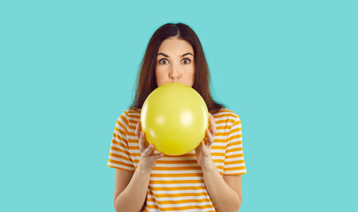 Woman blowing up birthday party balloon. Young brunette girl in striped orange white tee shirt isolated on turquoise background, inflating yellow balloon and looking at you with funny face expression © Studio Romantic