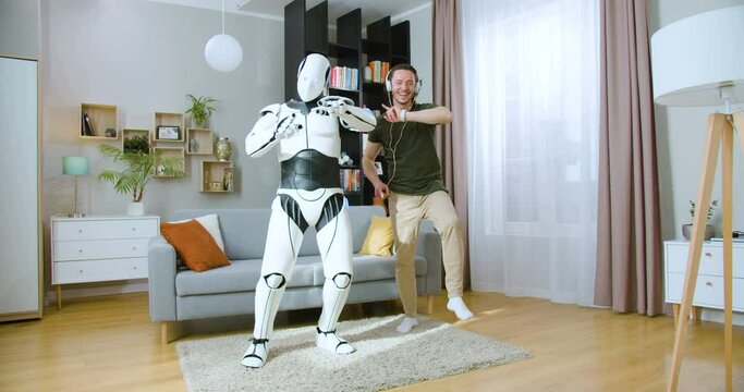 Human like futuristic robot and young caucasian male dancing together in apartment. Carefree happy man in white headphones having fun while listening music at home.