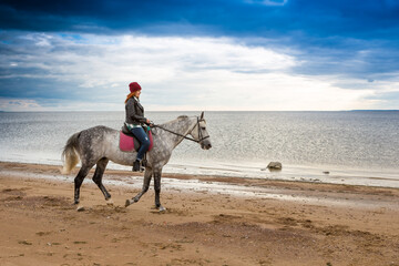 dressing jacket and jeans red-haired female horseback rider gallops on a dappled palfrey along sandy sea shore