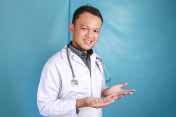 Happy young Asian man doctor, medical professional is smiling and pointing at a copy space isolated over blue background
