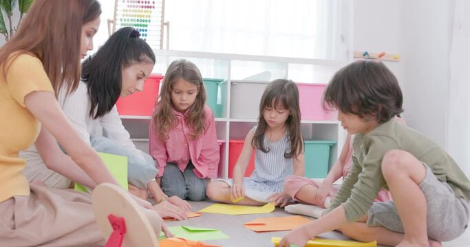 Children paint on drawing paper while sitting at the table next to their kindergarten teacher, who also draws something on the girl's paper while talking to the kids.