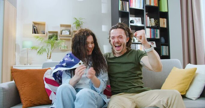 Front view of young man and woman covered with American flag sitting on sofa and watching sport TV. Happy sport fans celebrating victory of favorite team during competition.