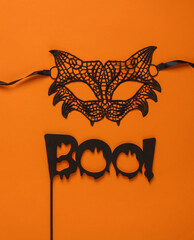 carnival lace cat mask and the word boo on a stick, orange background. Halloween party. Top view