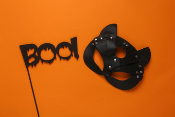 BDSM leather cat mask and the word boo on a stick, orange background. Halloween party