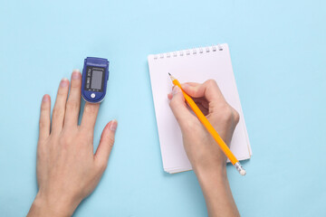 Hand finger with a pulse oximeter and hand writing data in a notebook on a blue pastel background....