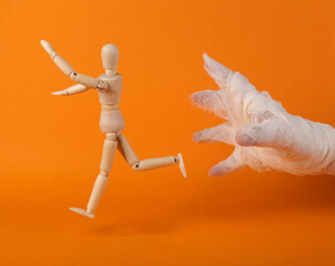 Frightened Wooden puppet runs away from the hand of a mummy wrapped in white bandages on an orange...
