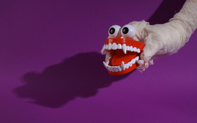 Mummy's hand holds monster's jaw with fangs, clockwork toy on purple background with shadow