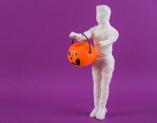 Trick or treat. Mummy doll wrapped in bandages with a jack-o'-lantern candy bucket on a purple background. Halloween minimal still life. Creative layout